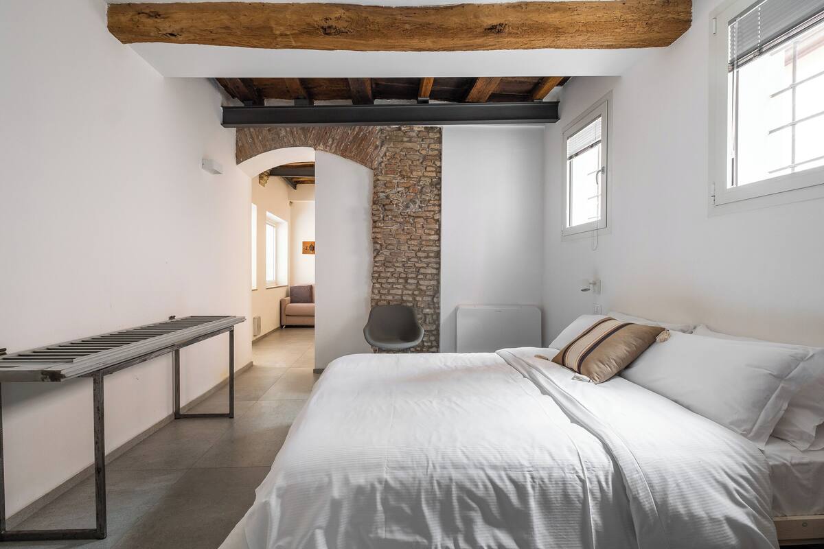 Un dormitorio con cama, un banco, una silla y una pared de piedra.