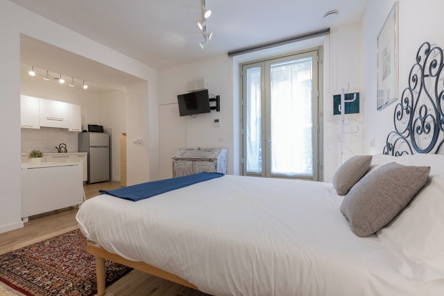 Un dormitorio con cama, almohadas, una colcha azul, piso de madera, una zona de cocina con armarios y electrodomésticos, y un televisor en la pared.