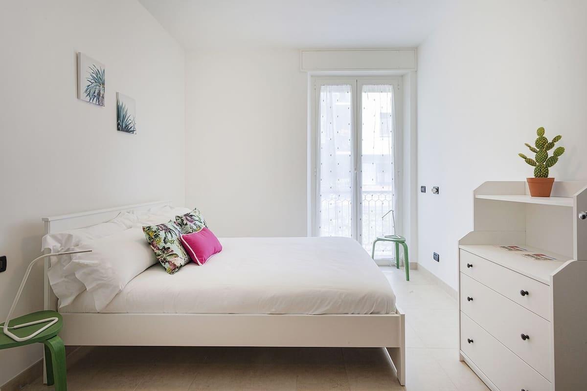 A minimalist bedroom with a single bed, a green chair, a dresser, and a potted cactus.
