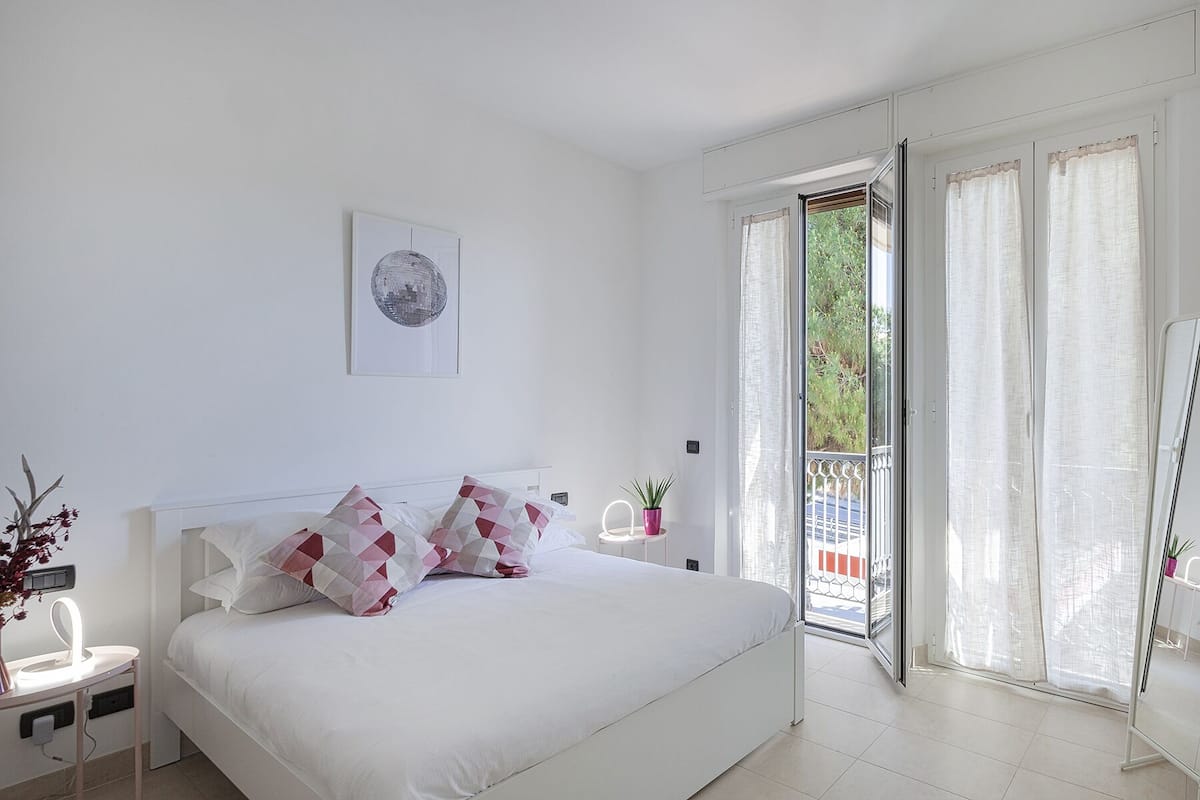 A bedroom with a bed, a bedside table, a potted plant, and a view of the outside through a glass door.