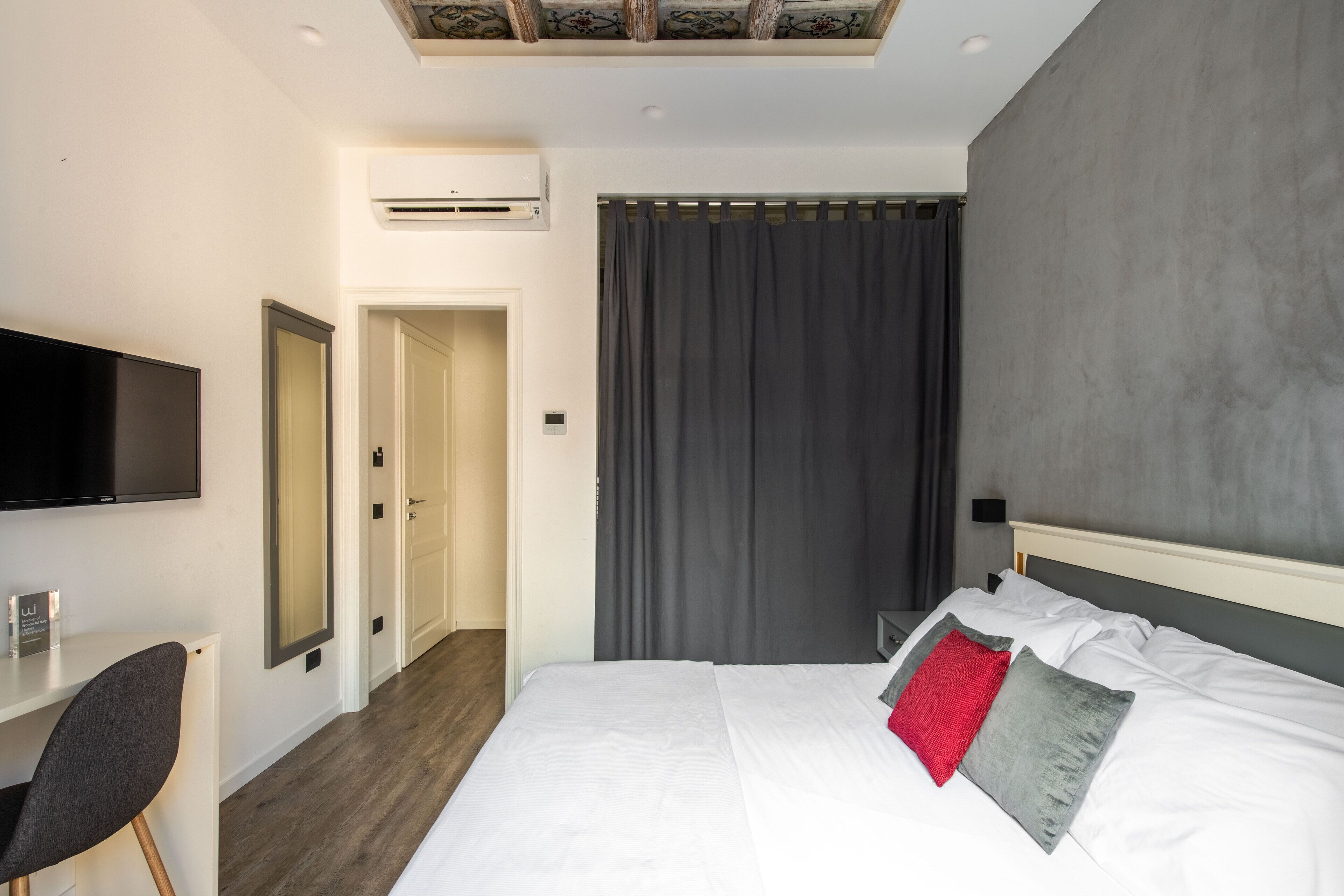 A modern hotel room with a large bed, a flat-screen TV mounted on the wall, a desk, and a door leading to another room.