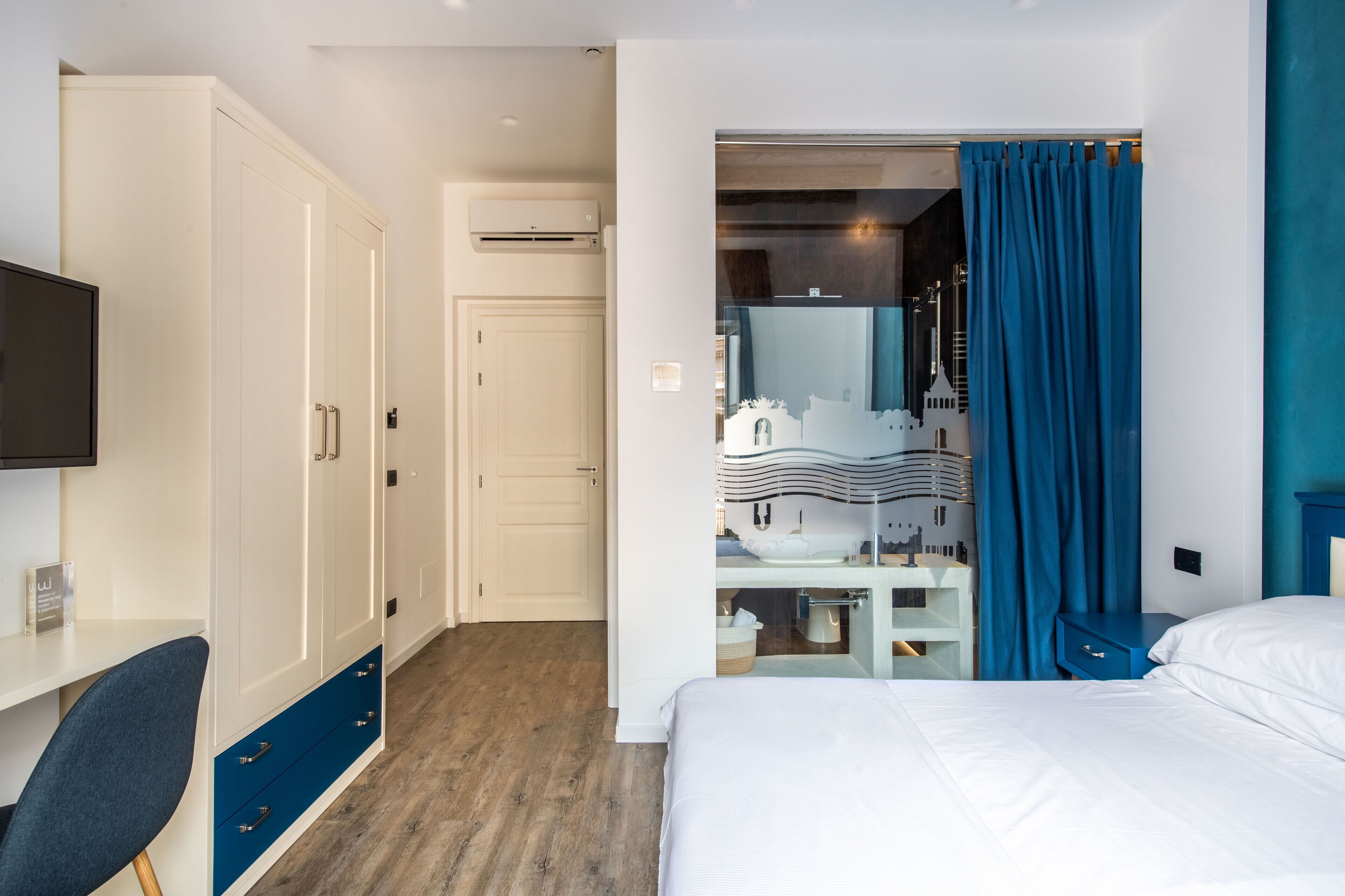 Habitación de hotel con cama, escritorio con silla, armario y baño visible a través de una puerta abierta.