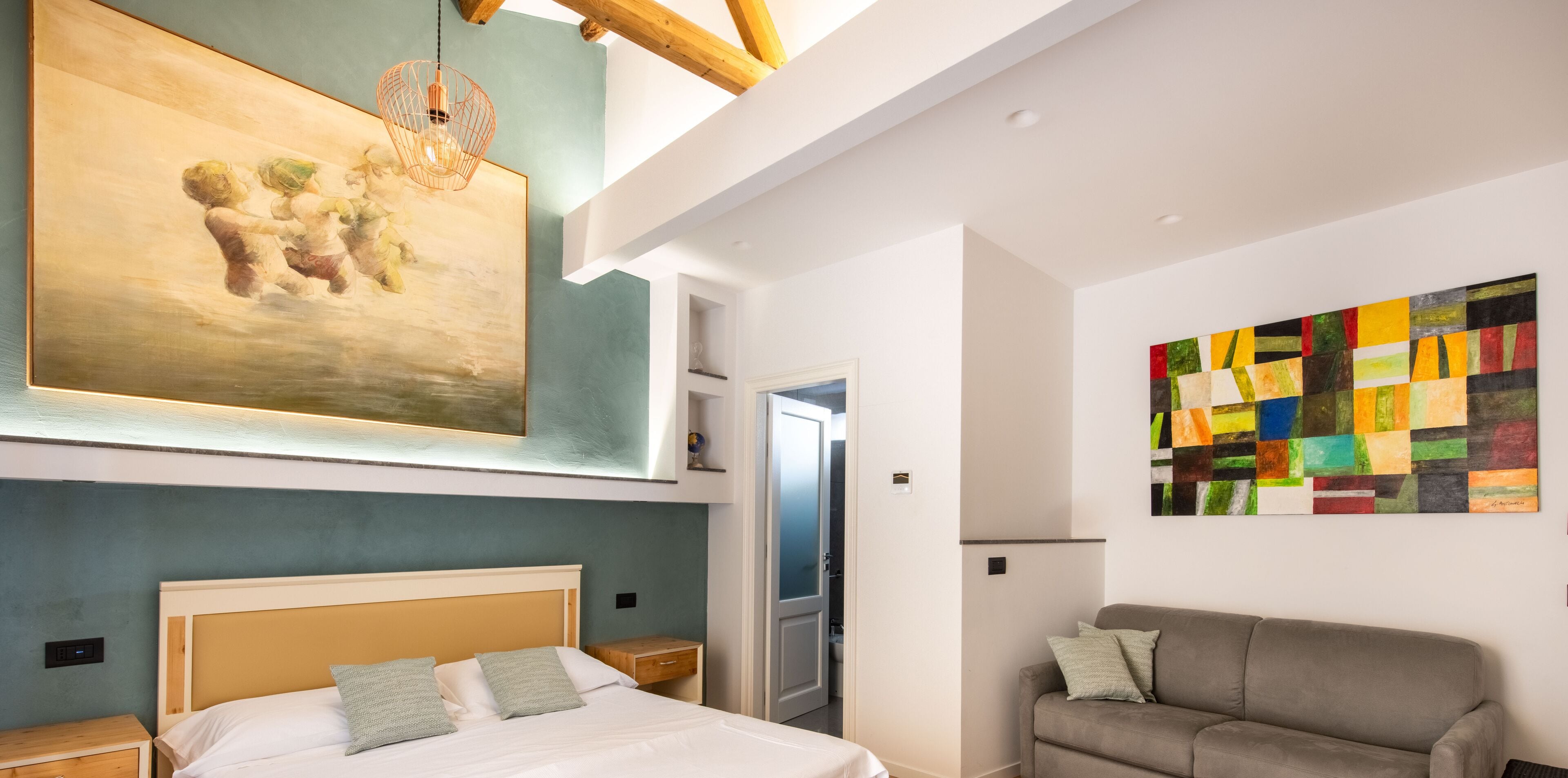Una habitación de hotel moderna con una cama, un sofá gris, un cuadro abstracto de colores y una viga de techo de madera.