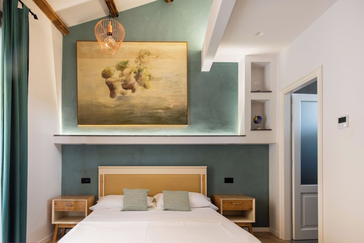 A modern bedroom with a large bed, bedside tables, a painting on the wall, and a view of a door.
