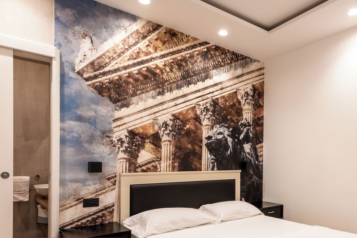 A modern bedroom with a bed, a nightstand, a wall-mounted lamp, and a large mirror.