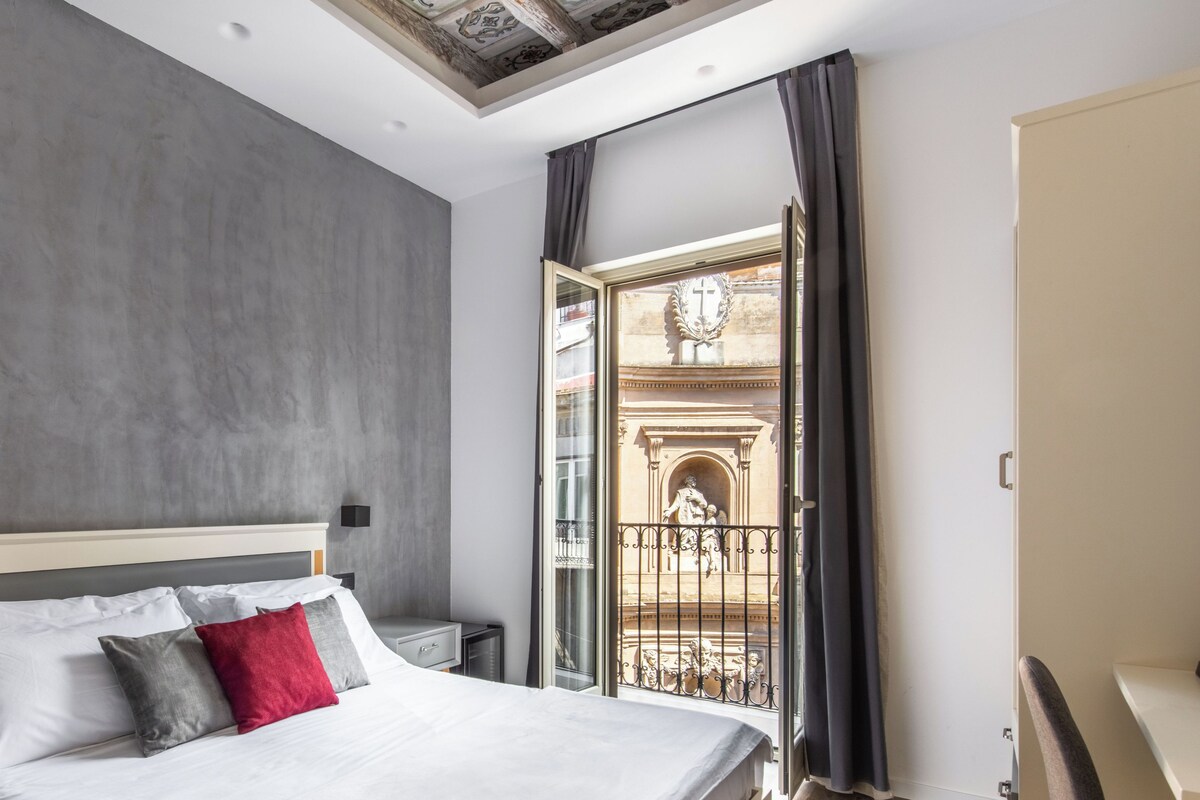 A modern bedroom with a large bed, a desk, and a balcony with a wrought iron railing.