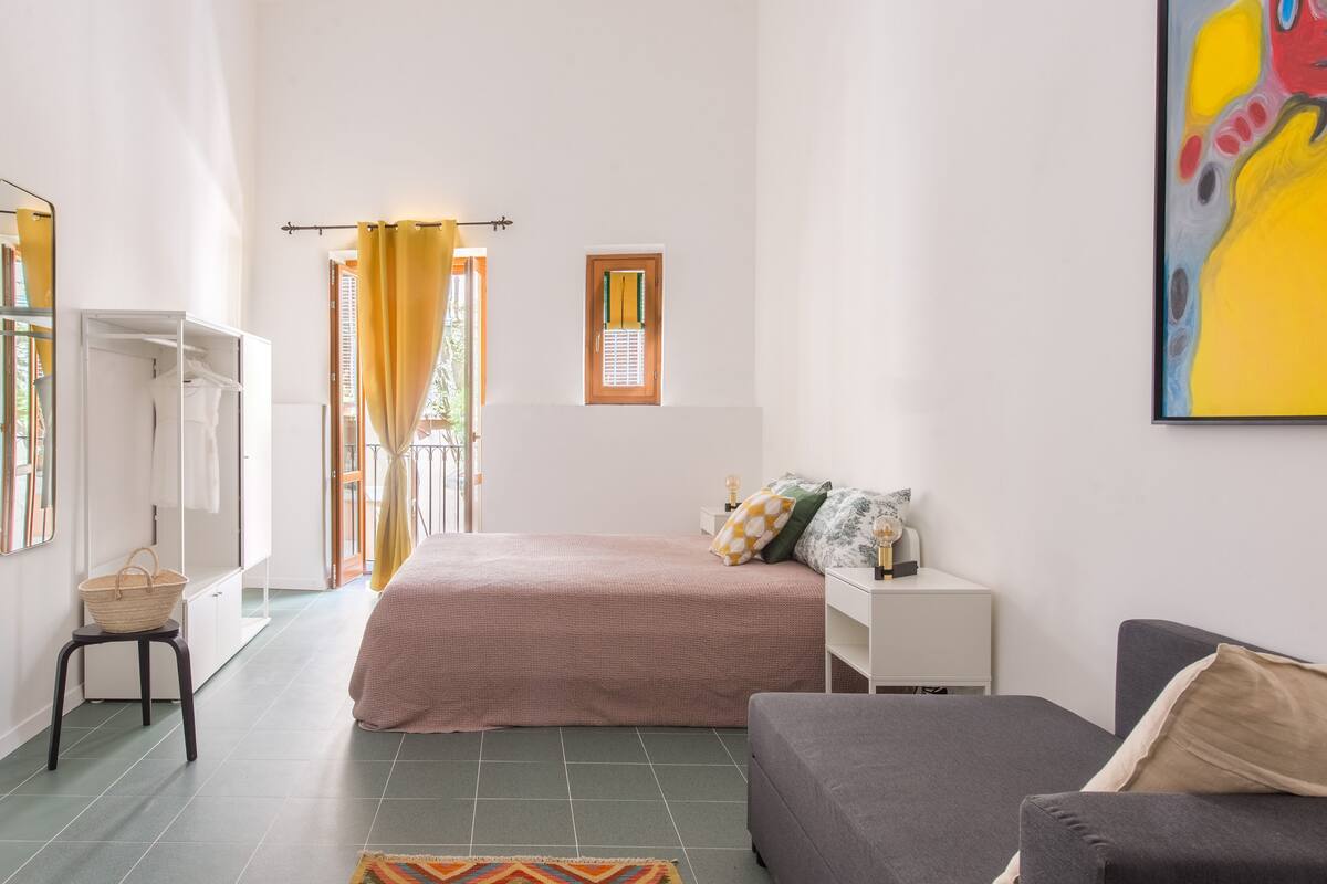 A bedroom with a bed, a sofa, a nightstand, a window with curtains, a small table with a basket, and a painting on the wall.