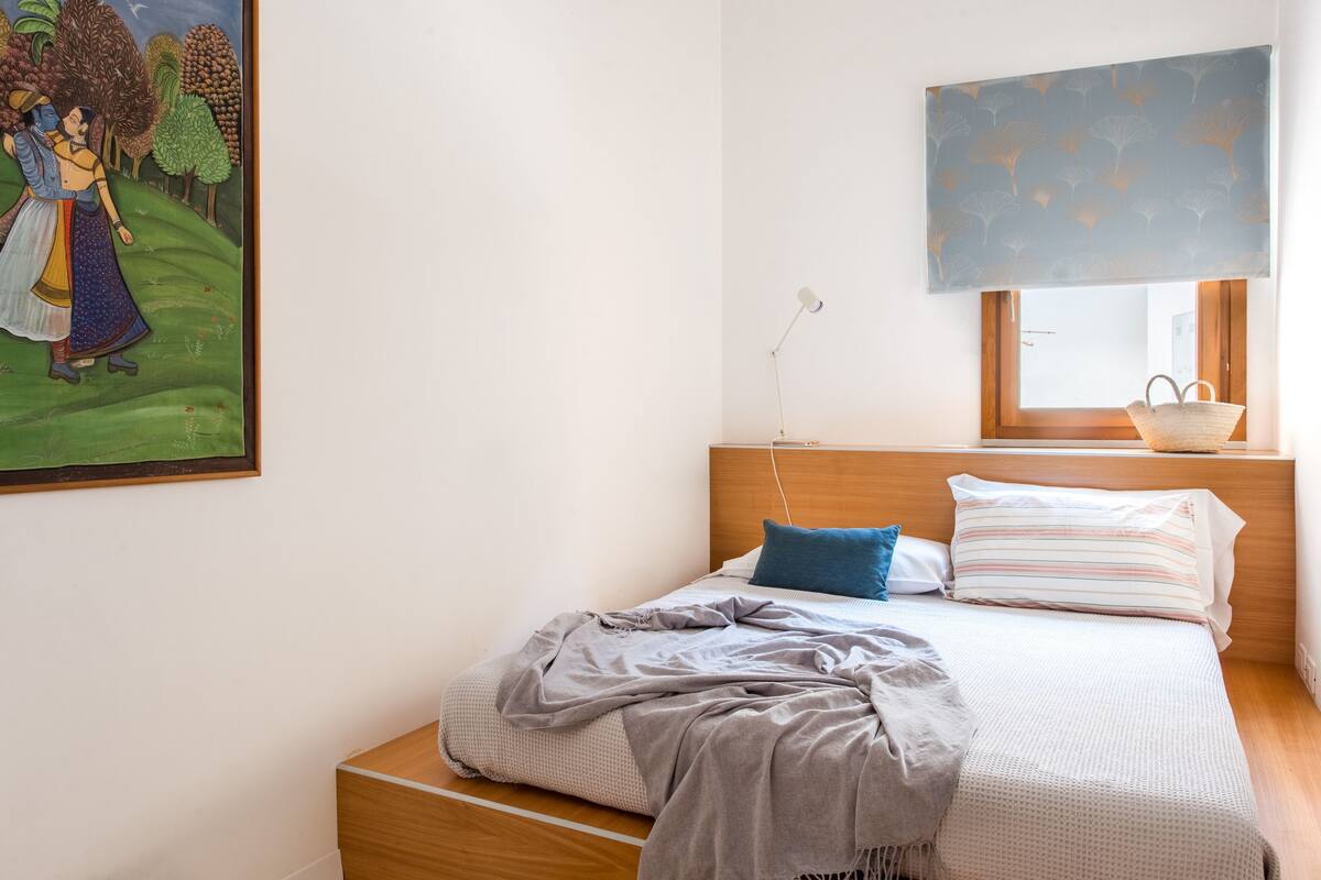 A bedroom with a bed, a nightstand, a lamp, a window with a view, and a painting on the wall.