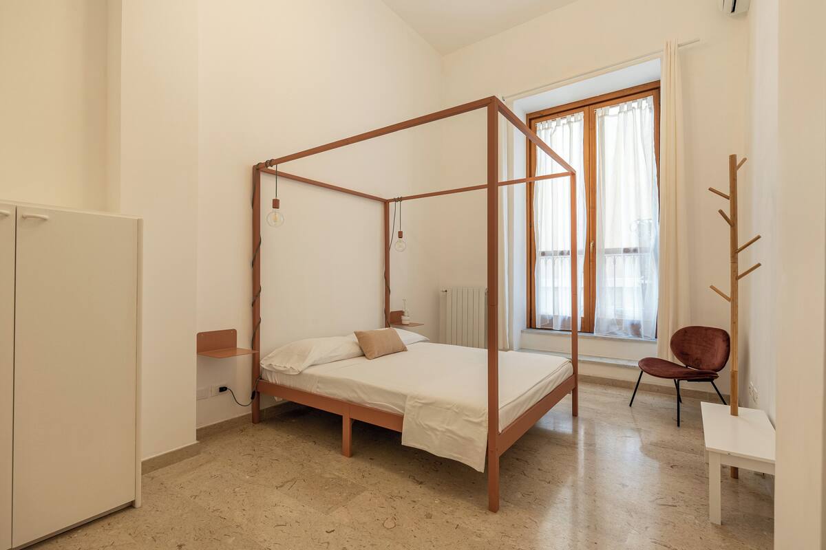 A bedroom with a four-poster bed, a chair, a small table, and a window with curtains.