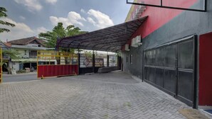 A paved courtyard with a covered area, a red gate, and a building with a large garage door. - Capital O 3198 Sukomanunggal Inn (Surabaya)