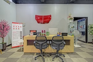 Un mostrador de recepción con dos sillas de oficina, un objeto rojo montado en la pared, una pizarra y una planta en maceta.