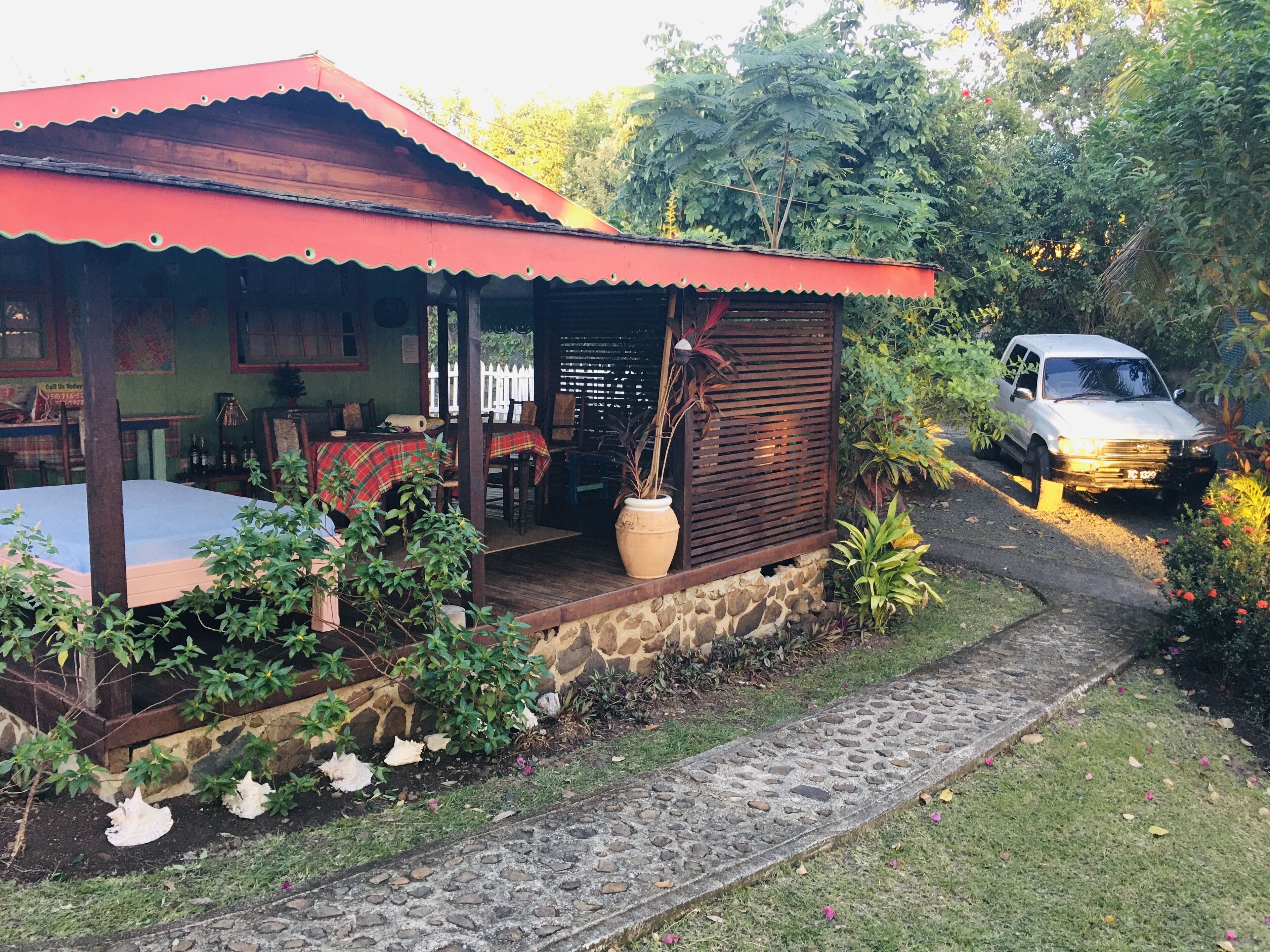Una casa de madera rústica con techo rojo, un porche con mesa y sillas, y un jardín con plantas y un camino.