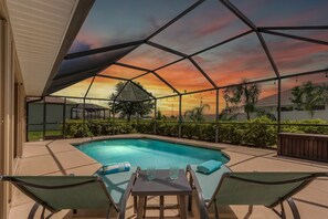 Piscina exterior techada con sillones de descanso y una mesa, rodeada de vegetación y vistas al atardecer.