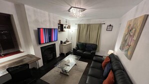 Ett modernt vardagsrum med en svart lädersoffa, ett soffbord i glas, en öppen spis, en tv som är monterad på väggen och en kristallkrona.
