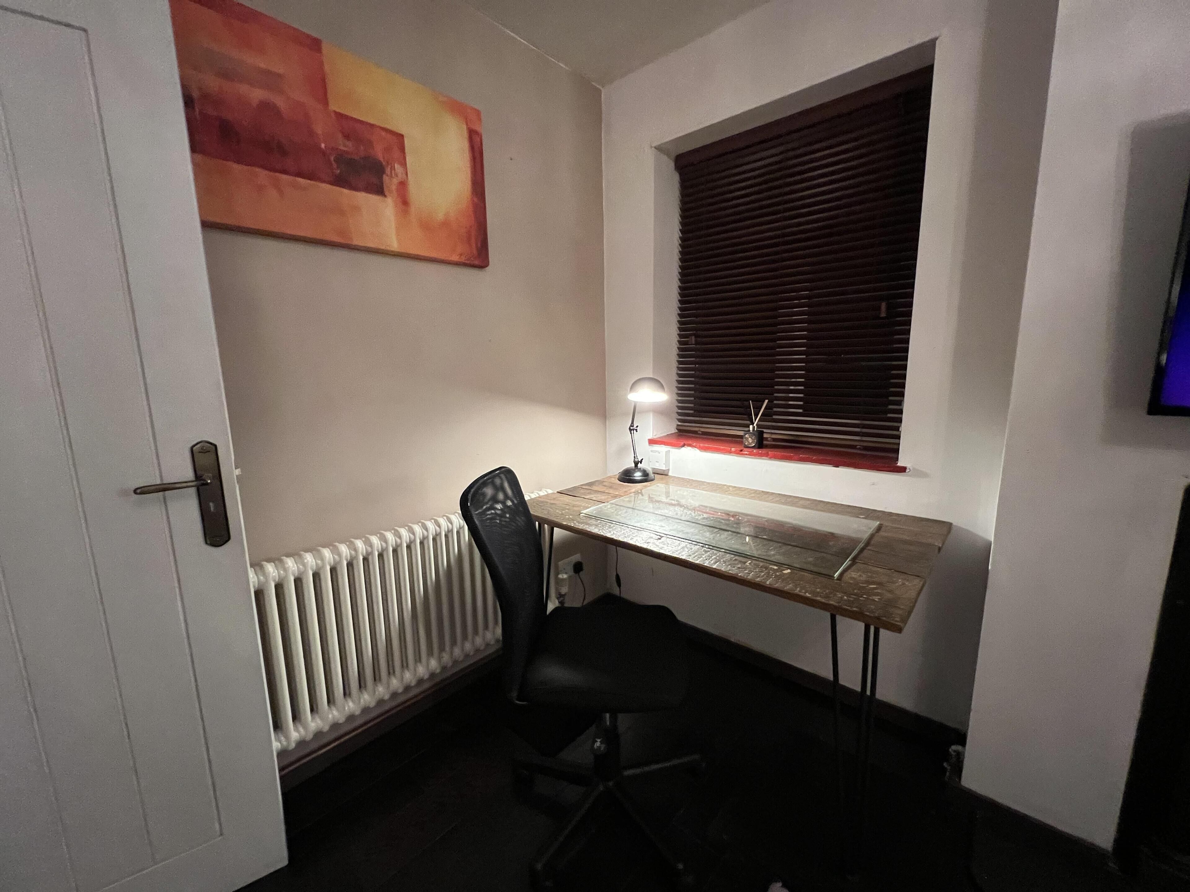 Ein kleiner, schlicht beleuchteter Raum mit einem Holztisch, einem schwarzen Bürostuhl, einem Heizkörper, einem Fenster mit Jalousien und einem Bild an der Wand.
