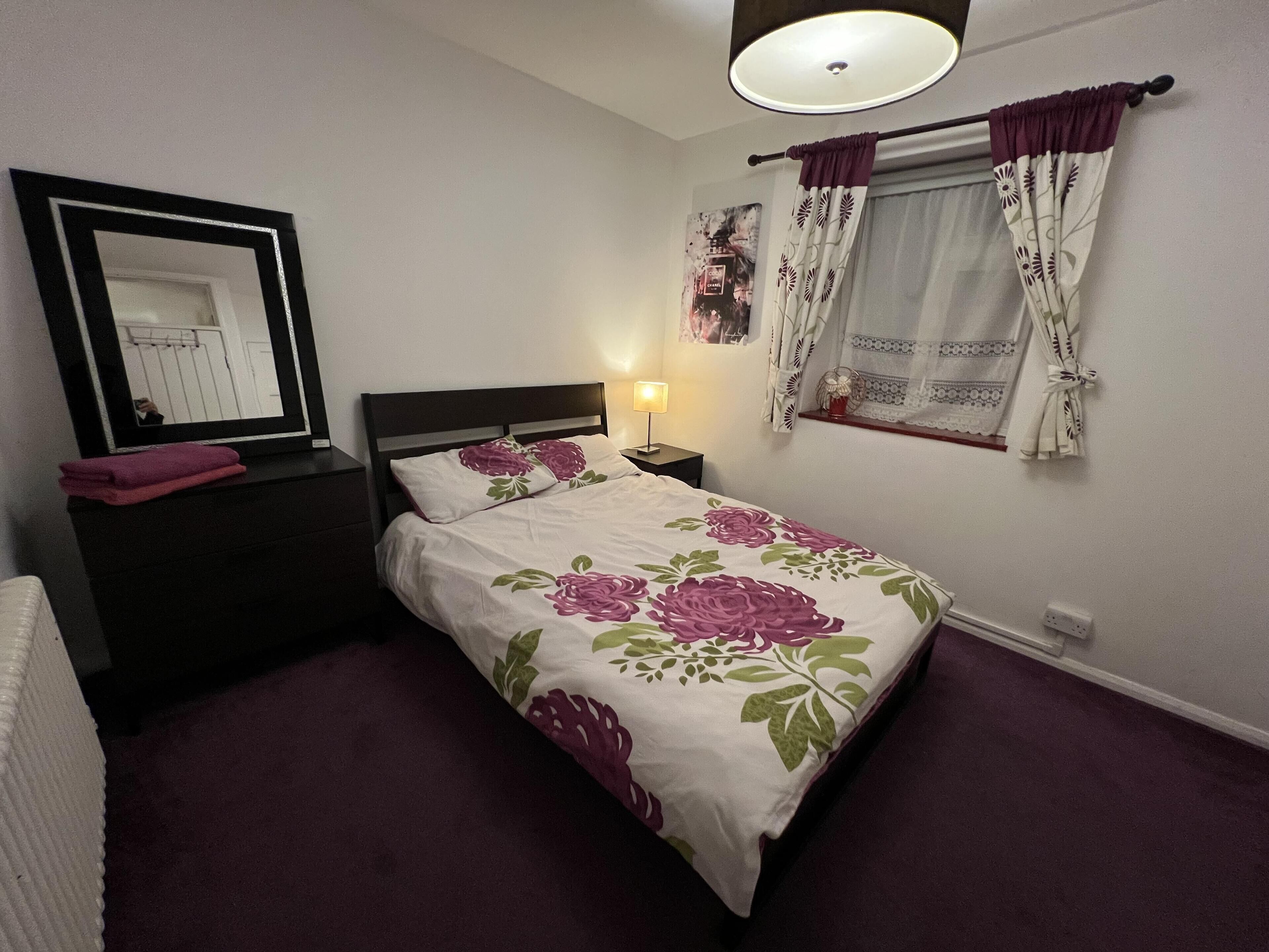 A bedroom with a bed, a nightstand, a mirror, a window with curtains, and a wall-mounted lamp.