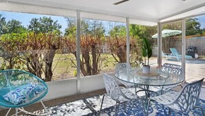 Un espace extérieur abrité, doté d’une table et de chaises en verre, d’un ventilateur de plafond et offrant une vue sur un jardin avec des arbres et une pelouse.