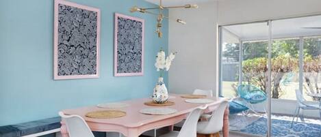 A dining area with a pink table, white chairs, and two framed artworks on the wall.