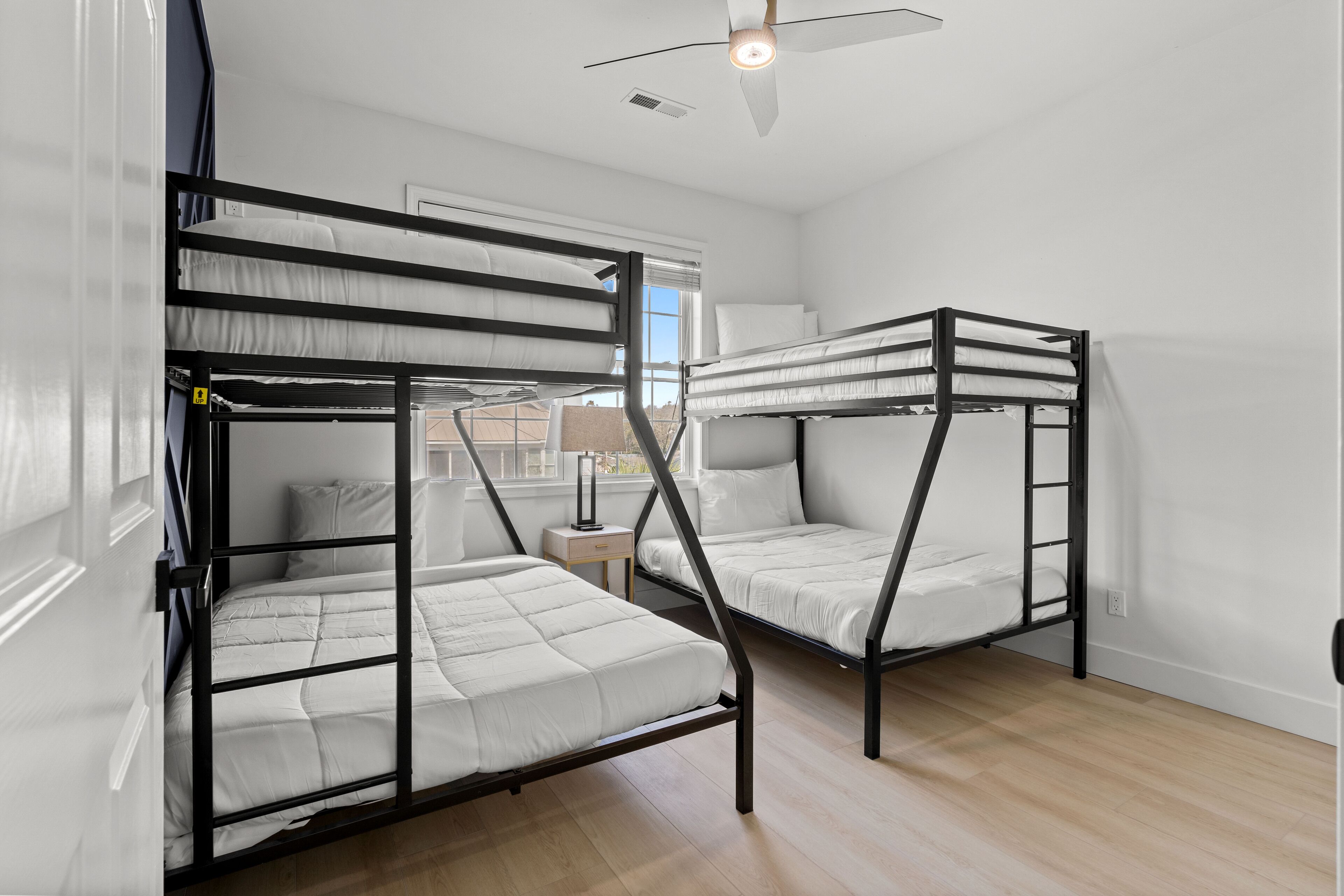 A room with two bunk beds, a small bedside table with a lamp, and a window with blinds.