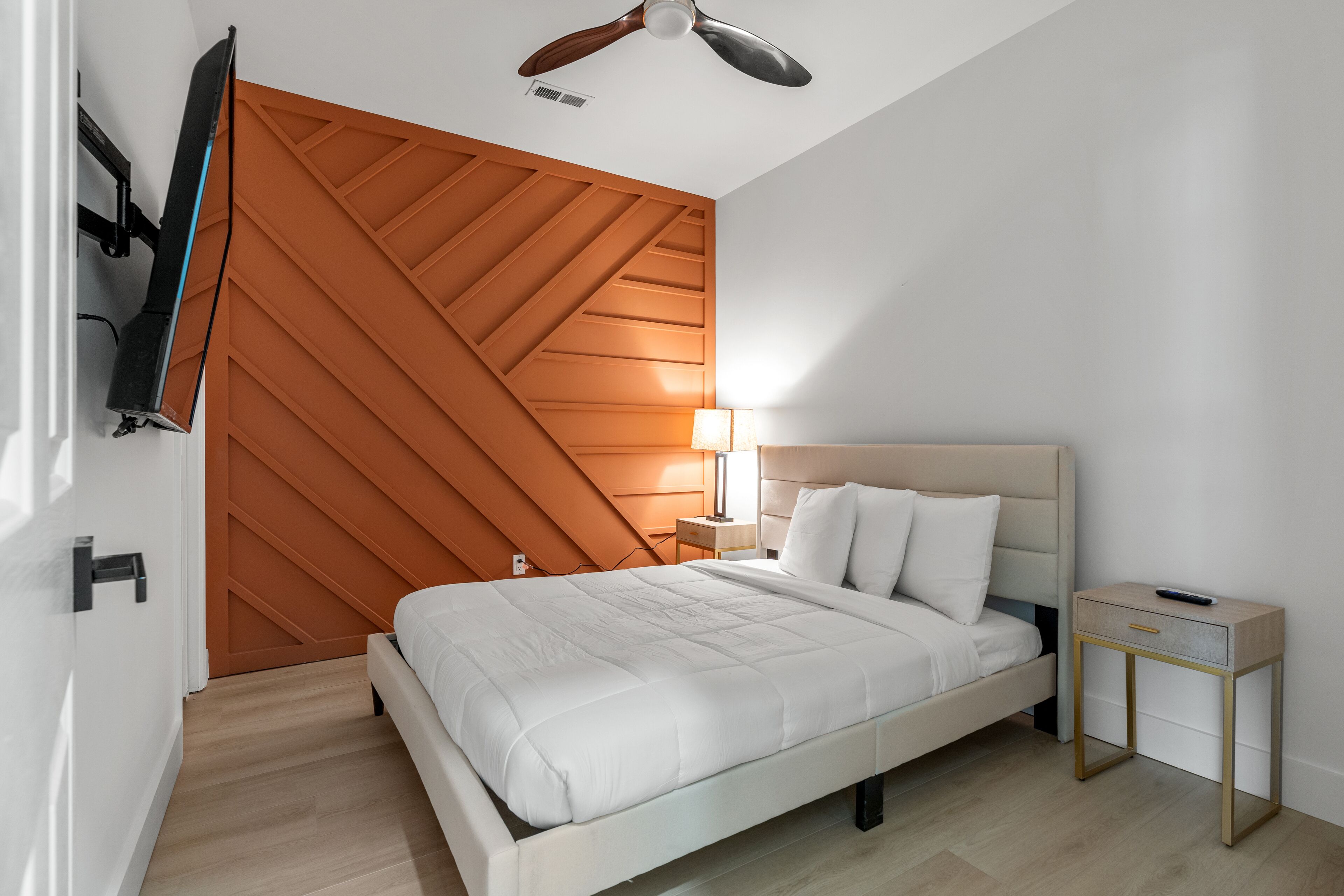 Un dormitorio moderno con cama, mesita de noche, televisor montado en la pared y ventilador de techo.