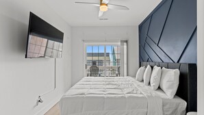 Une chambre moderne avec un lit, un ventilateur de plafond, un téléviseur fixé au mur et une vue sur l’extérieur par une fenêtre.