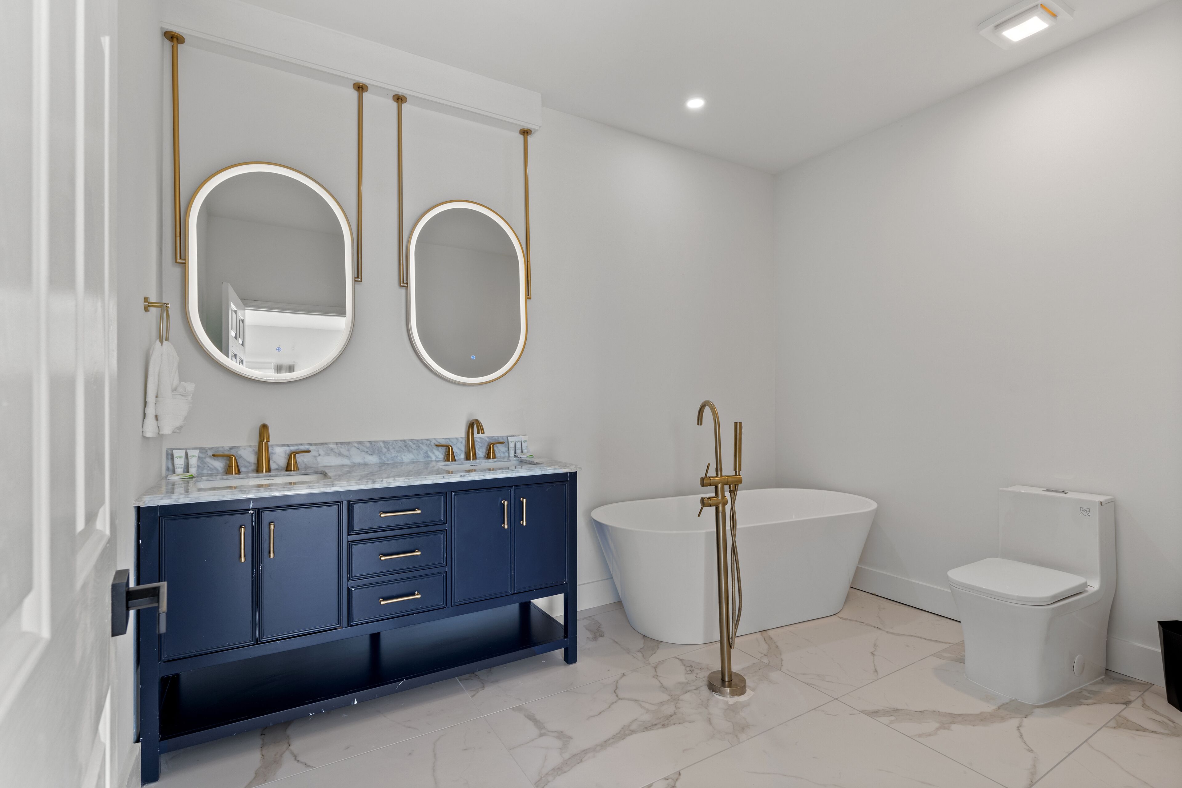 Une salle de bain moderne avec un meuble-lavabo bleu foncé, deux miroirs ovales, une baignoire sur pieds et des toilettes.