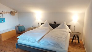 Una cama doble con ropa de cama blanca, dos mesitas de noche, un perchero en la pared, una pequeña mesa redonda y suelo de madera.