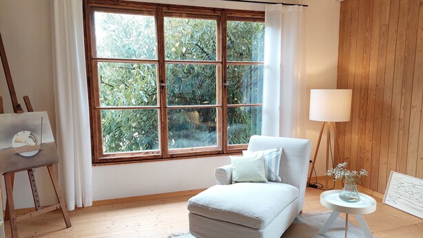 Una habitación con un ventanal amplio, un sillón blanco, una mesita con una lámpara y un caballete con una pintura.