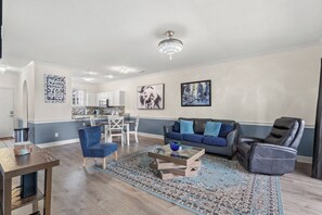 Una sala moderna con sofá, sillón y una mesa de centro, un comedor con sillas y una cocina al fondo.