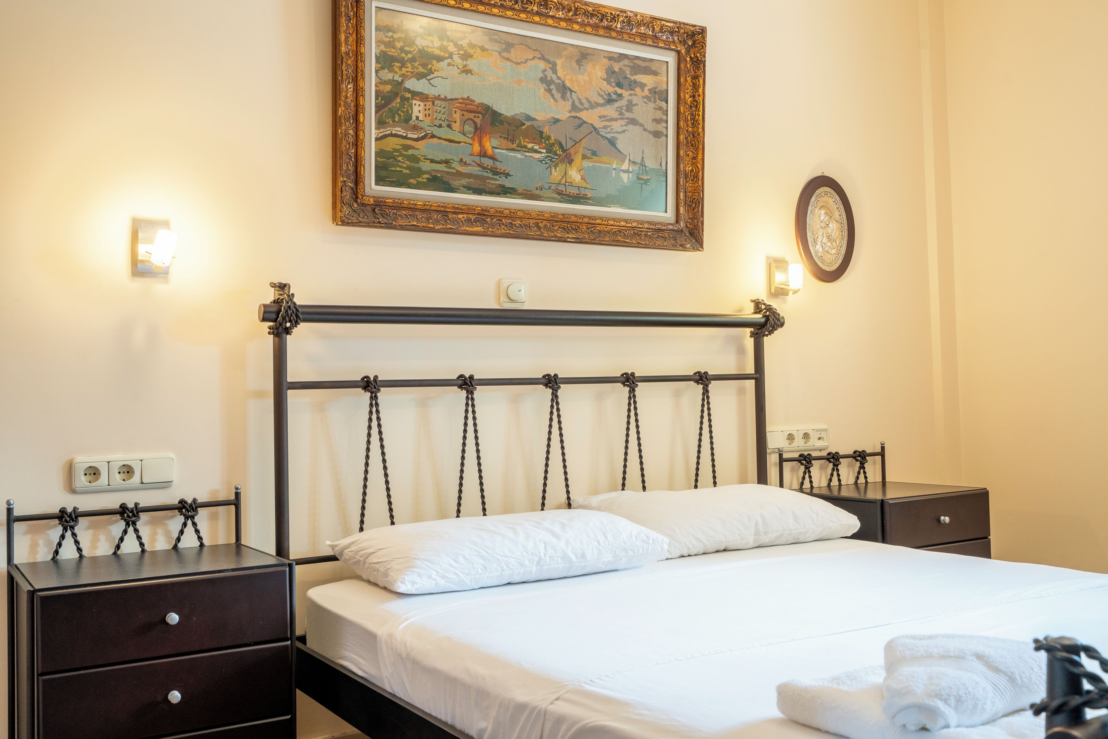 A hotel room with a bed, two bedside tables, a nightstand, a wall-mounted painting, and a wall socket.