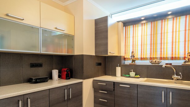 A modern kitchen with dark cabinets, a countertop, a sink, and a window with striped curtains. - Seaside Luxury Residence near the Airport (Nea Makri)