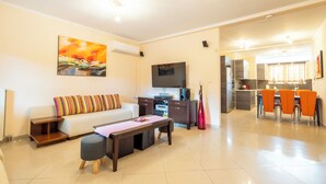 A living room with a sofa, coffee table, and TV, a dining area with chairs, and a kitchen in the background. - Seaside Luxury Residence near the Airport (Nea Makri)