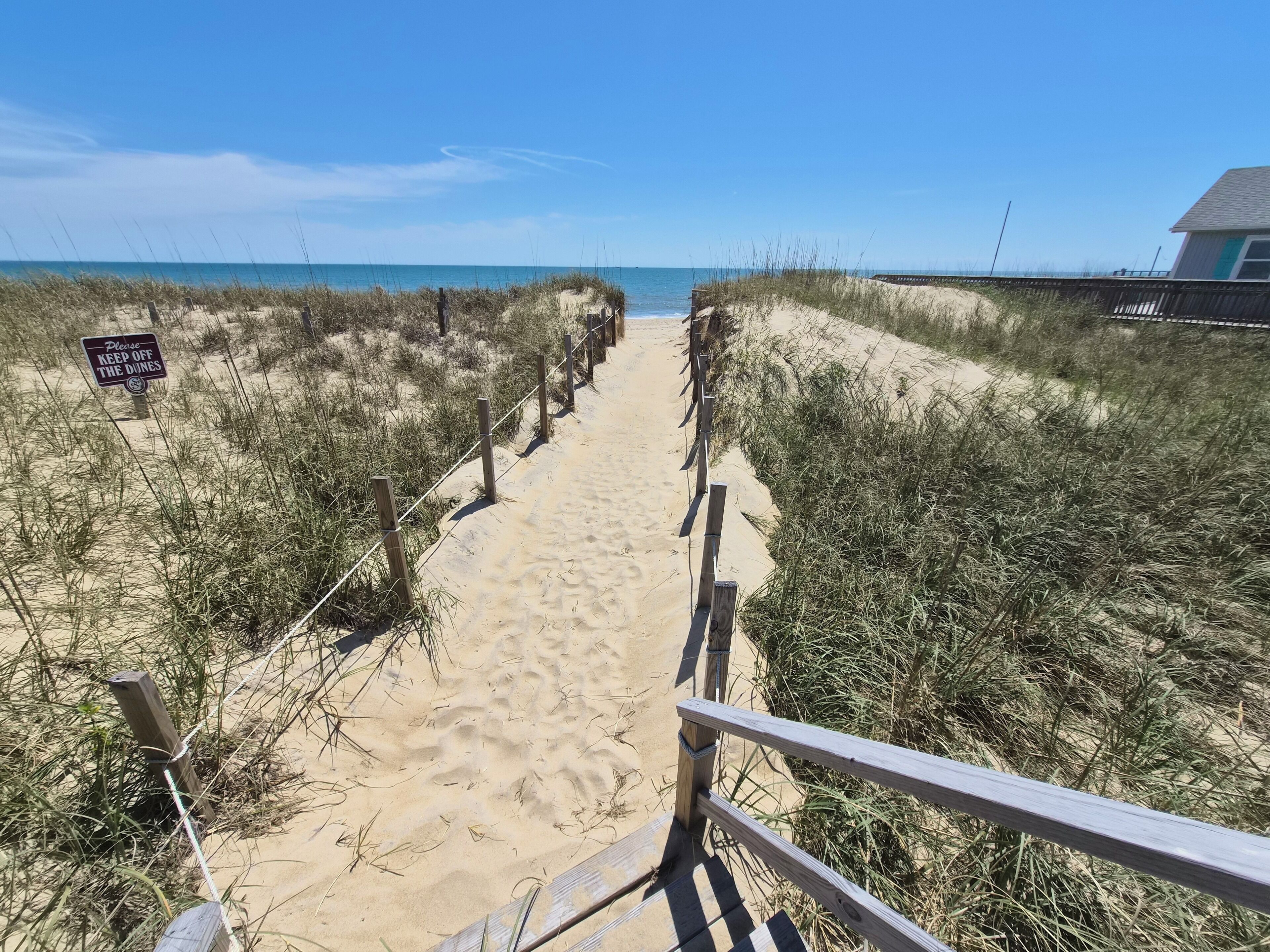 A sandy beach path leading to the ocean, flanked by wooden fences and a sign.