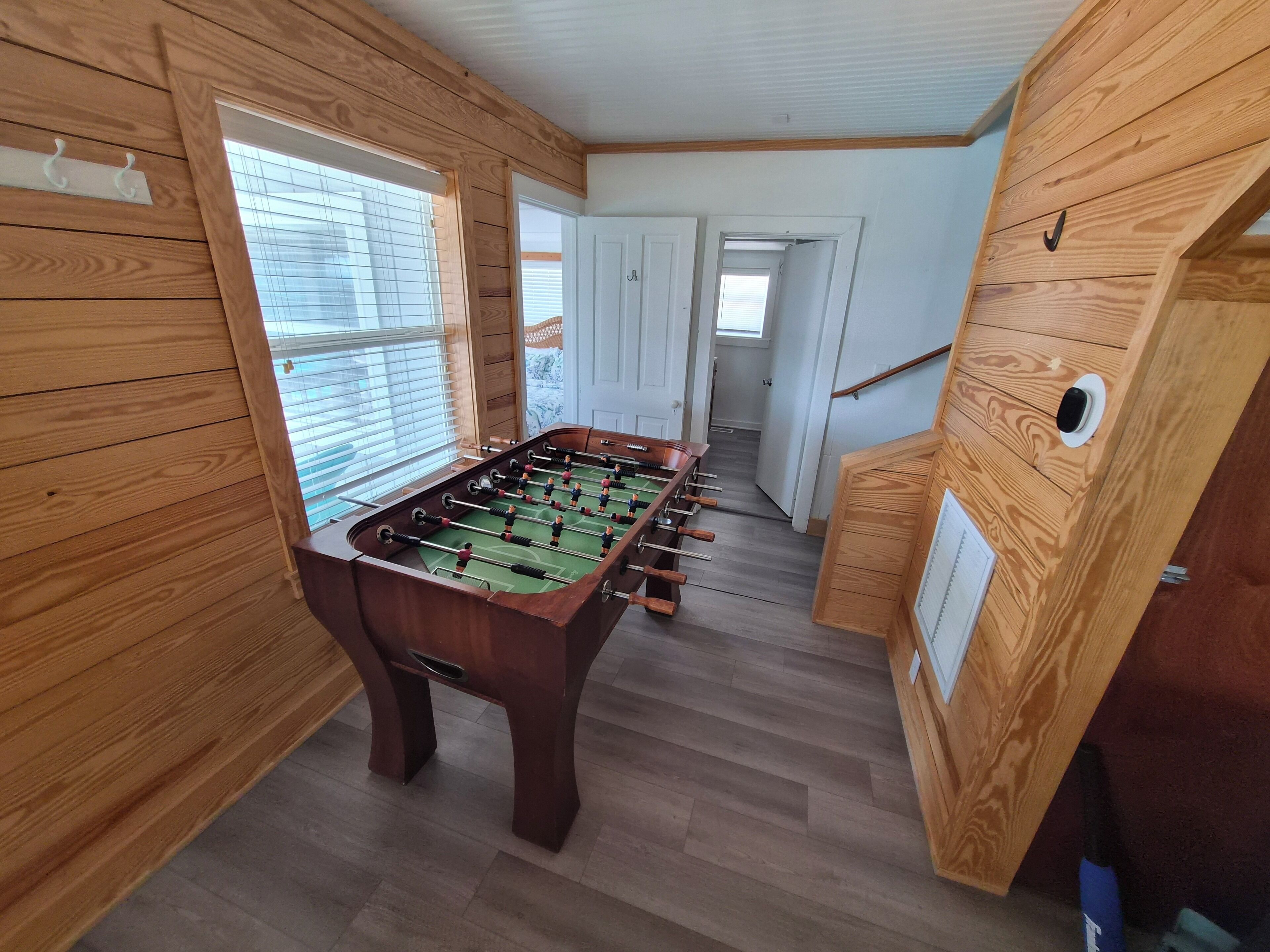 Um quarto com piso de madeira, mesa de pebolim, paredes de madeira e porta que dá para outro cômodo.