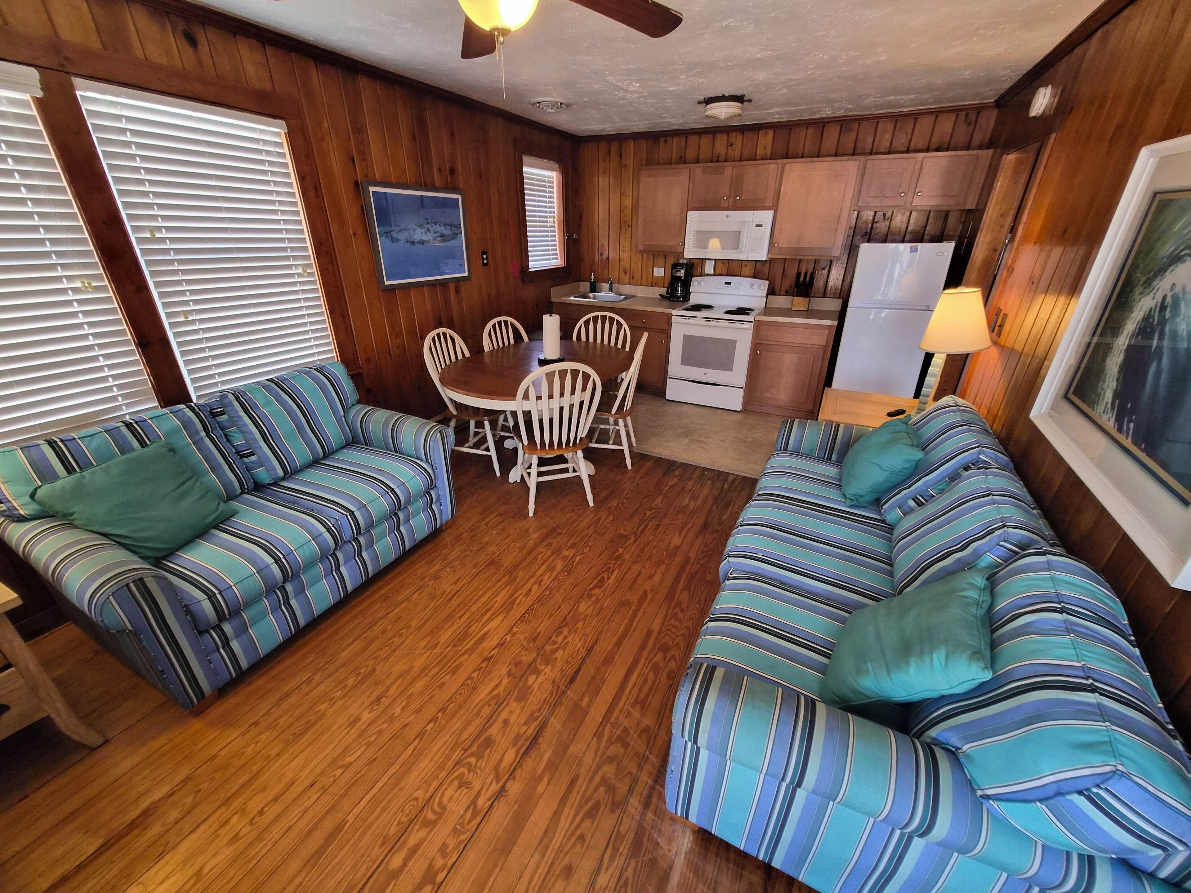 A cozy living room with striped sofas, a dining table, and a kitchen area with wooden cabinets and appliances.