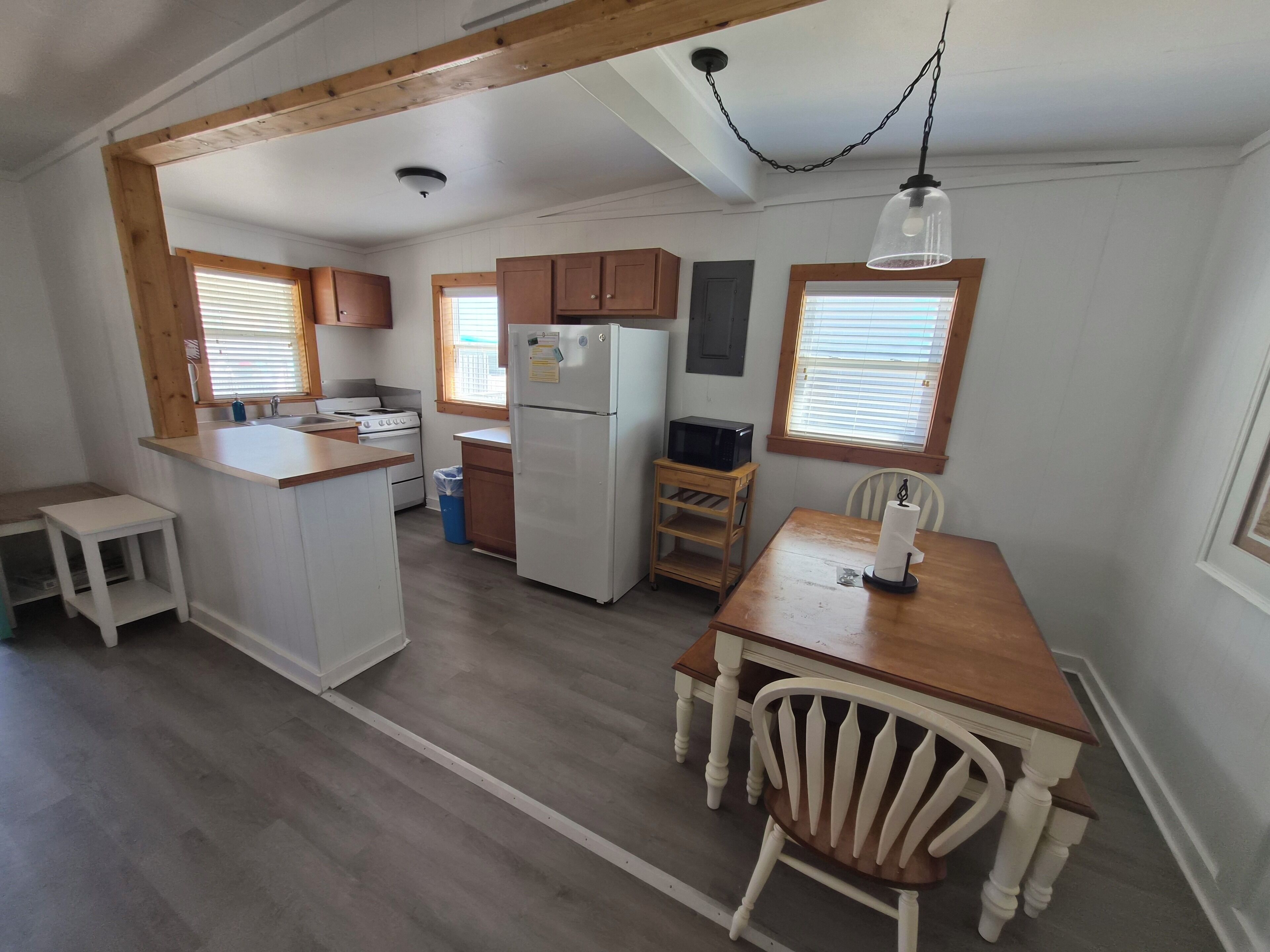 Una cocina con armarios de madera, un refrigerador, un microondas, una mesa de comedor de madera con una silla y una ventana con persianas.