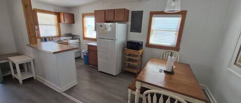 Una cocina con armarios de madera, un refrigerador, un microondas, una mesa de comedor de madera con una silla y una ventana con persianas.