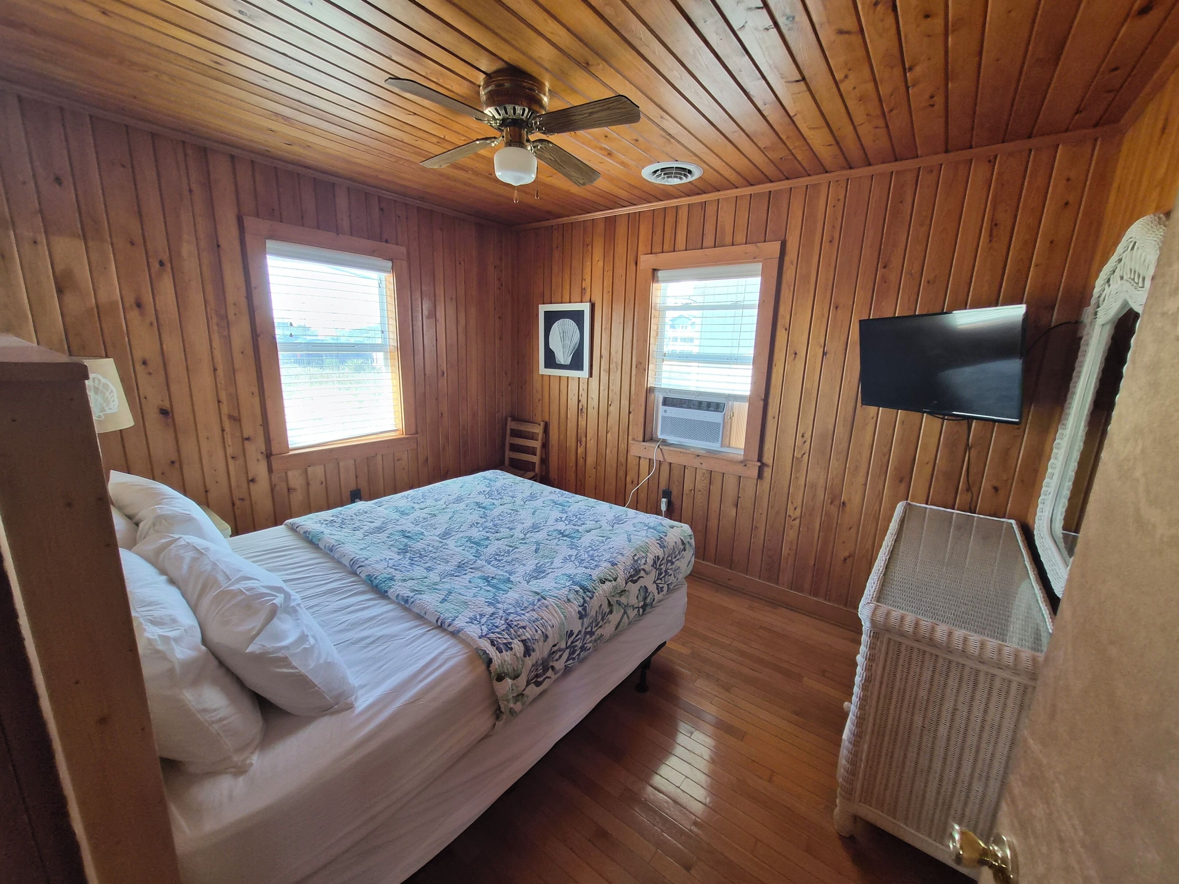 Ein Zimmer mit Holzwänden, einem Bett, einem Deckenventilator, einem Wand-Fernseher und einem Fenster.