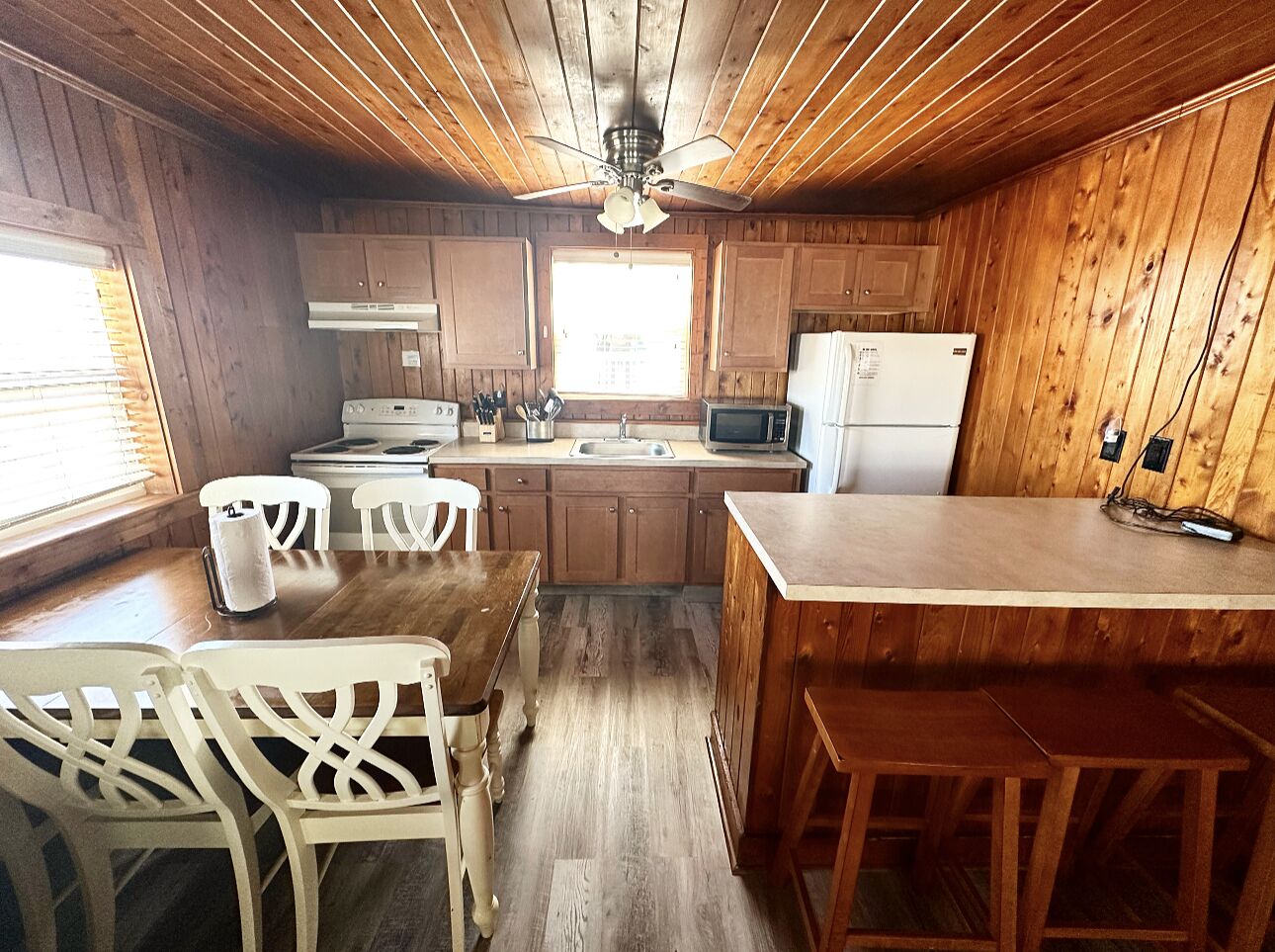 A wooden cabin-style kitchen with a dining table and chairs, a refrigerator, a microwave, and a ceiling fan.