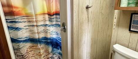 Une salle de bain avec un rideau de douche représentant un coucher de soleil sur l’océan, des toilettes et un meuble en bois contenant du papier toilette.