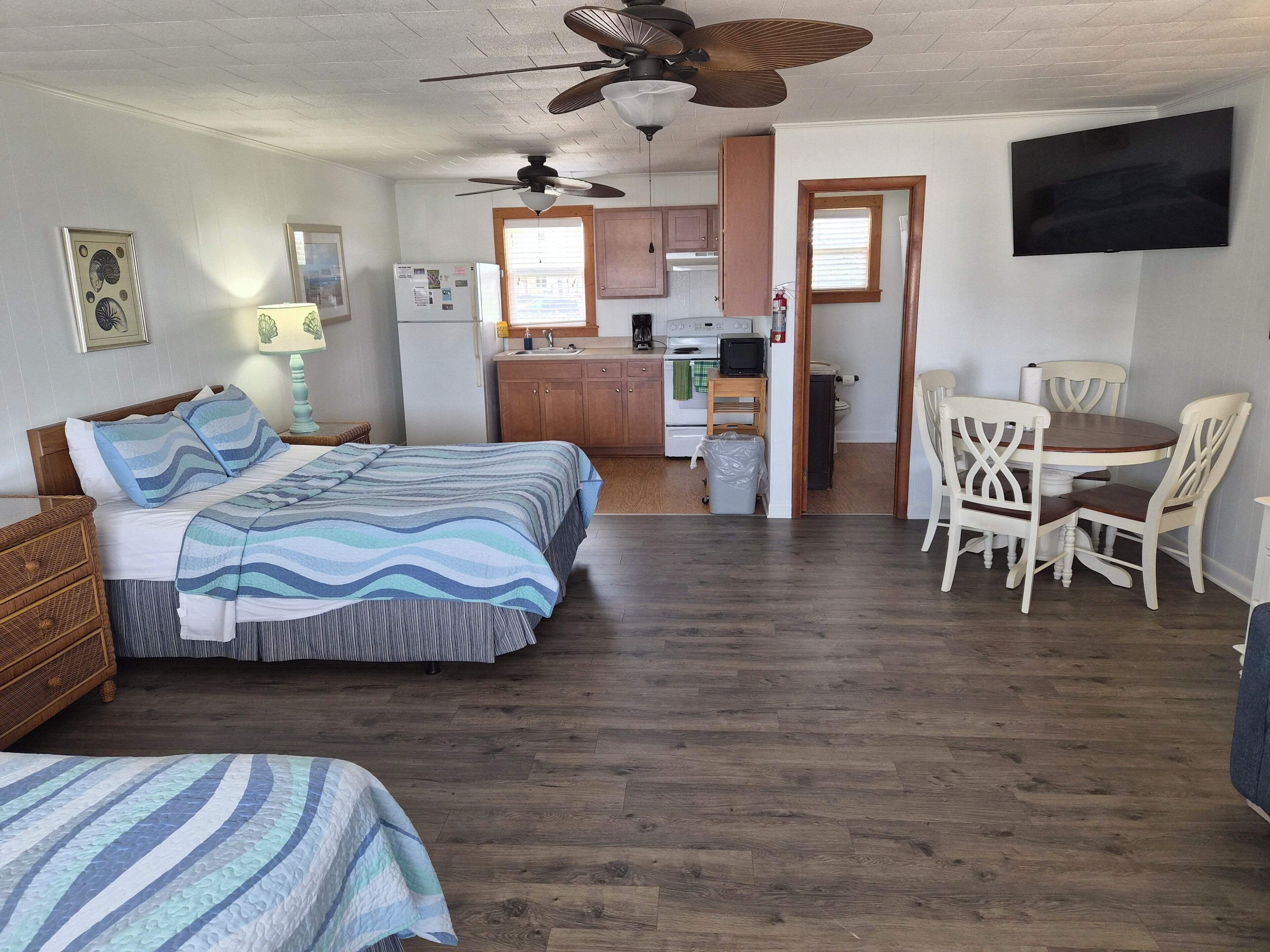 A bedroom with two beds, a dining area with chairs, a kitchen with cabinets, and a ceiling fan.