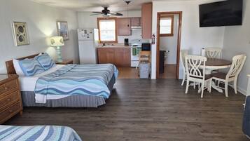 A bedroom with two beds, a dining area with chairs, a kitchen with cabinets, and a ceiling fan.