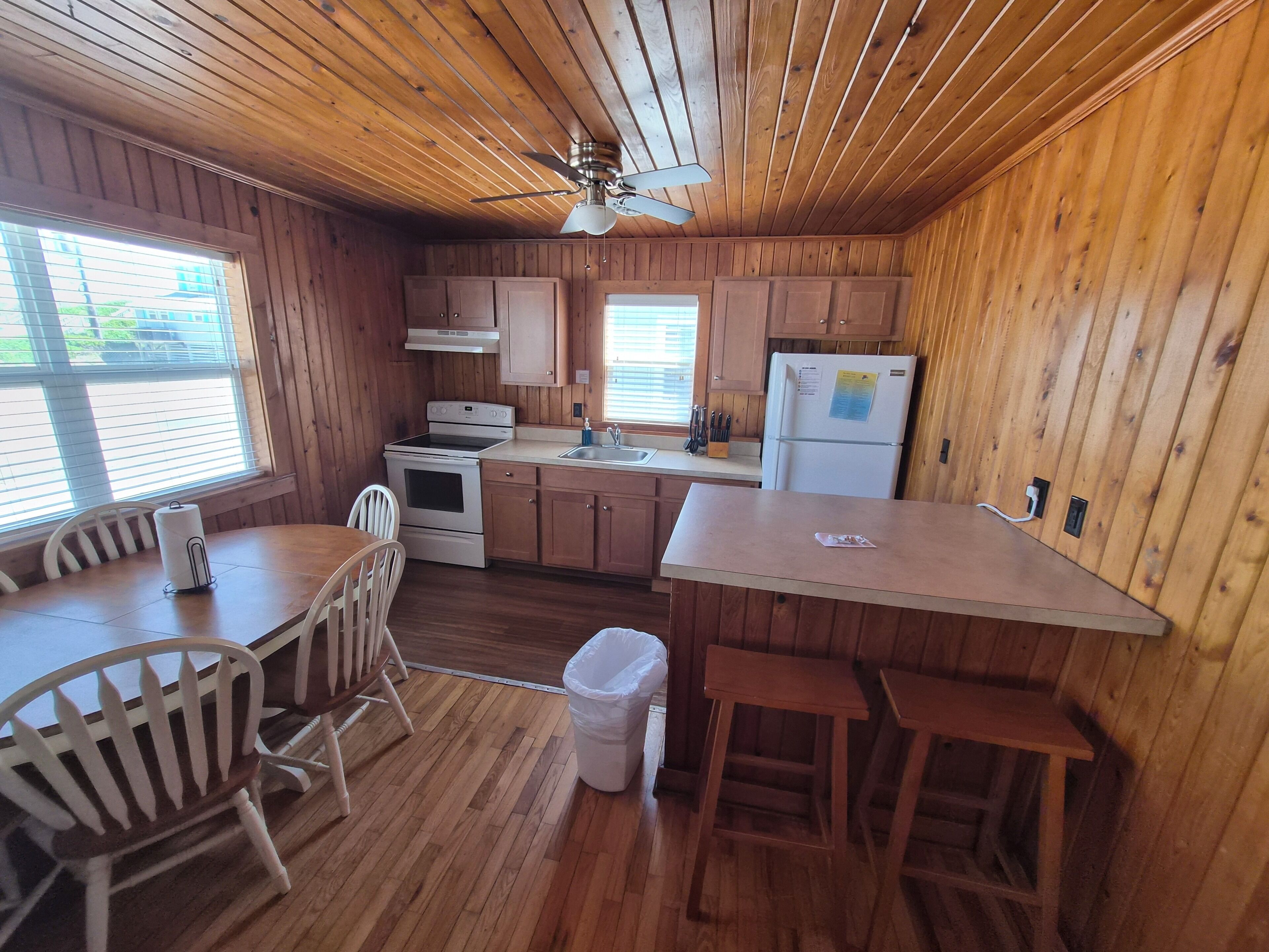 A wooden cabin kitchen with a dining table, chairs, a stove, oven, refrigerator, and a ceiling fan.