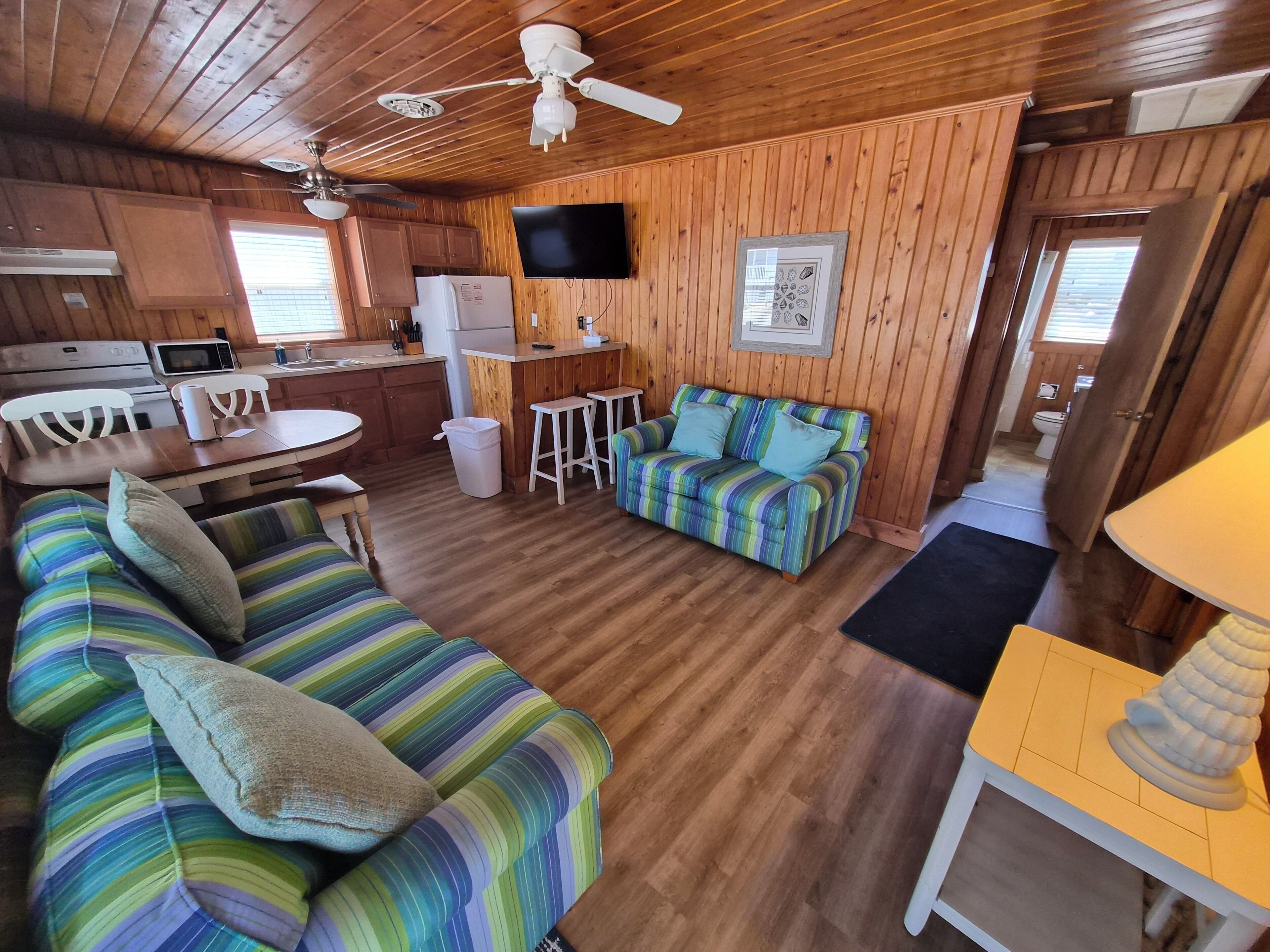 A cozy cabin interior with a wooden floor, a striped sofa, a dining table with chairs, a kitchen area, and a ceiling fan.