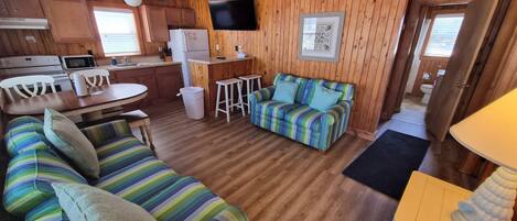 A cozy cabin interior with a wooden floor, a striped sofa, a dining table with chairs, a kitchen area, and a ceiling fan.