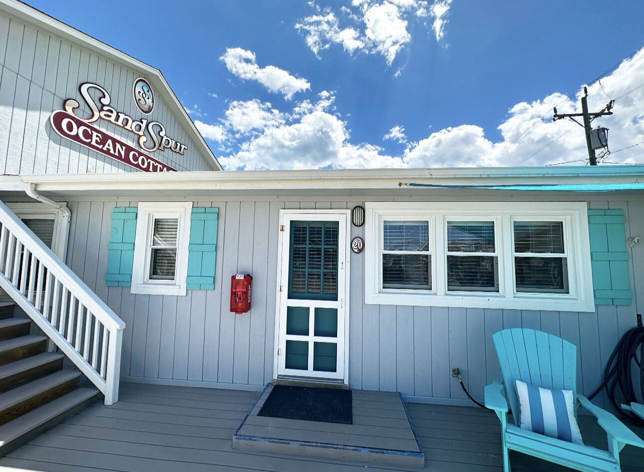 En kystbygning med skilt der siger "Sandspit Ocean Cottage". Den har en hoveddør, en trappe og en blå Adirondack-stol udenfor.