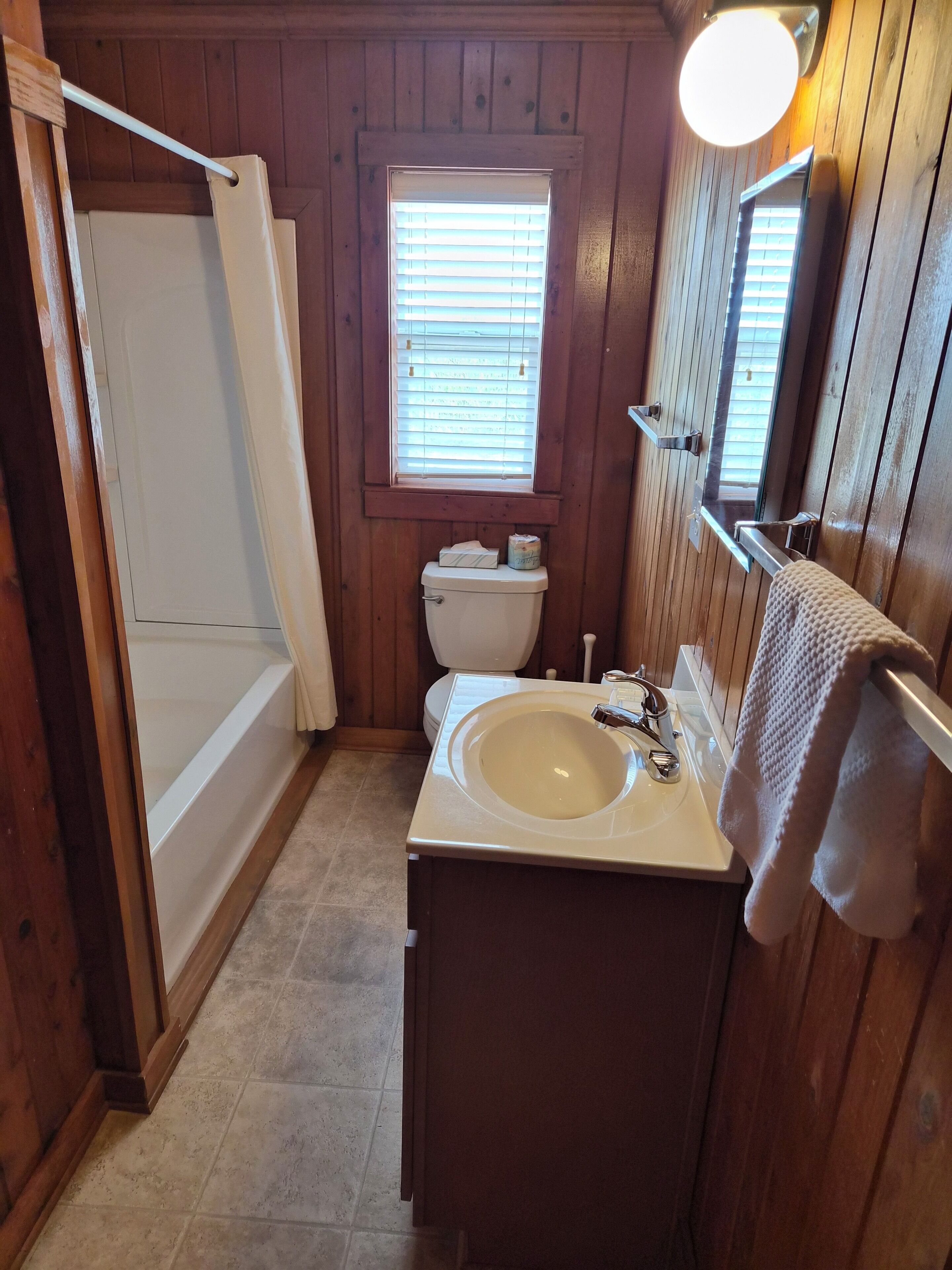 A wooden bathroom with a bathtub, toilet, sink, and a window with blinds.