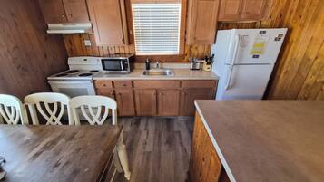 Una cocina compacta con muebles de madera, microondas, estufa, fregadero y refrigerador. Hay dos sillas blancas y una mesa de madera.