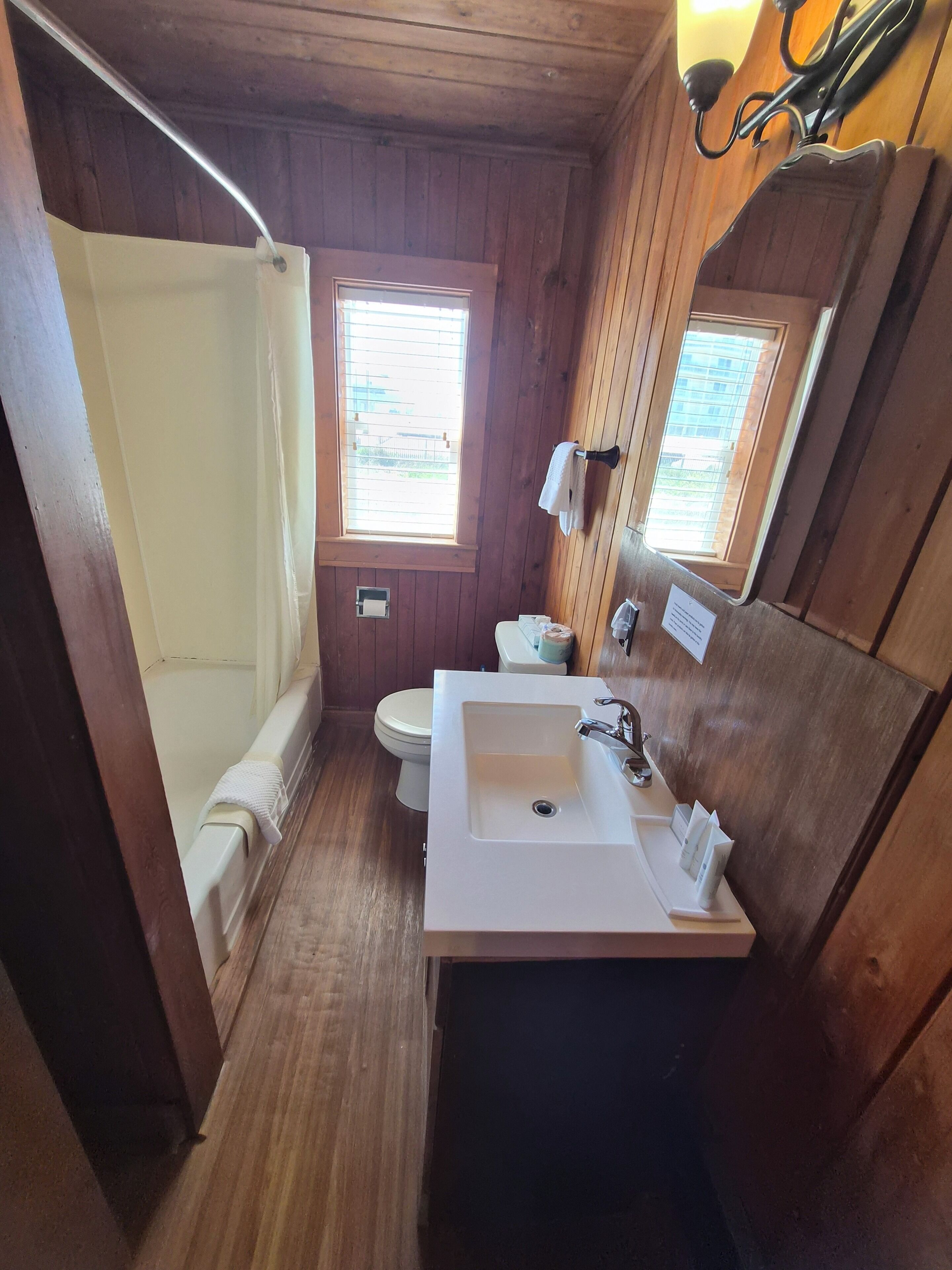 Een houten badkamer met een douche, toilet, wastafel en een raam.