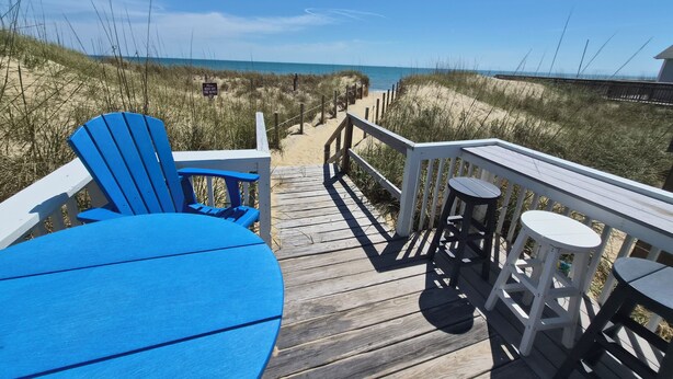 A wooden deck with a blue chair and table, a railing, and a view of the beach and dunes.